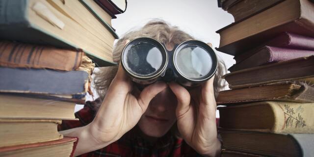 person hiding behind piles of books looking through binoculars